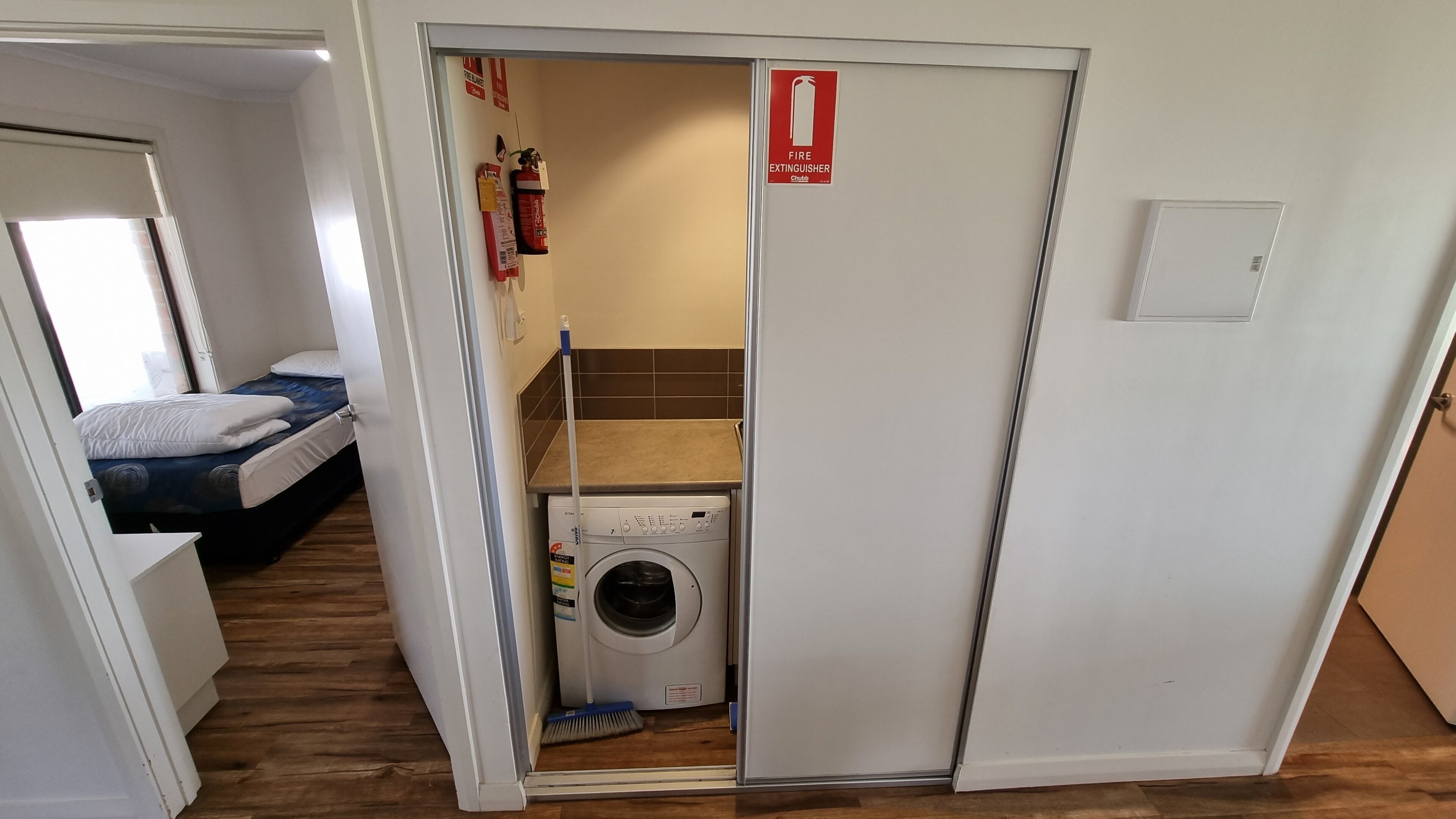 A compact room with a bed, a washing machine, and a small kitchenette area.