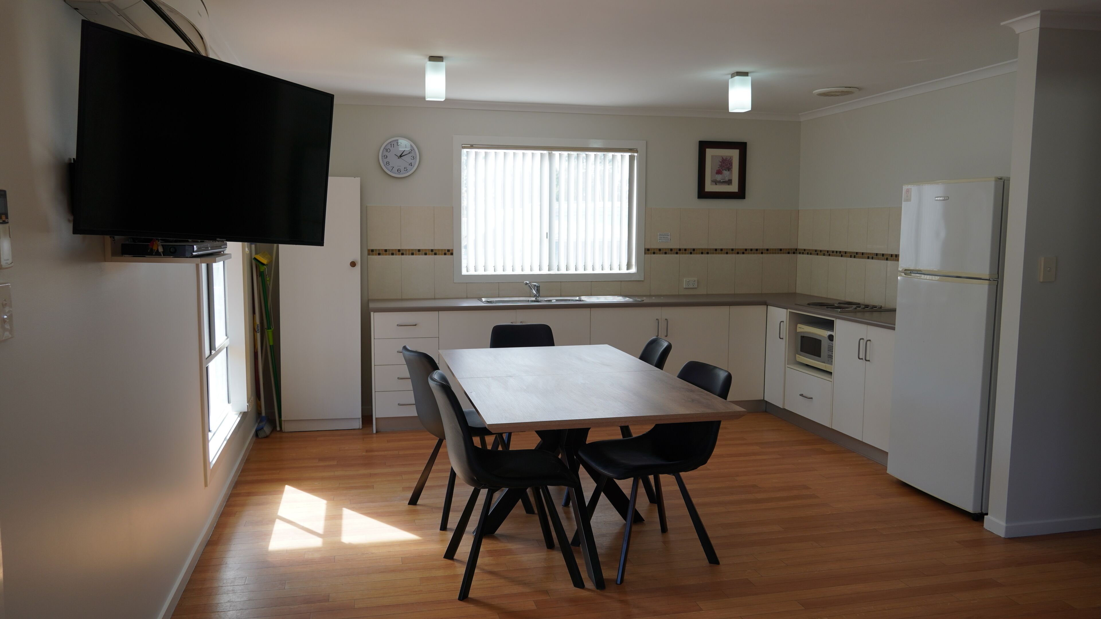 A compact kitchen with a dining table, chairs, a refrigerator, a microwave, and a TV mounted on the wall.