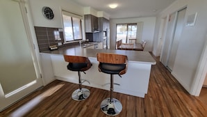 A modern kitchen with two bar stools, a dining area with a table and chairs, and a large window providing natural light. - Wallaroo Holiday Park (Wallaroo)
