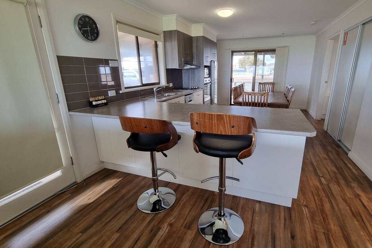 A modern kitchen with two bar stools, a dining area with a table and chairs, and a large window providing natural light.