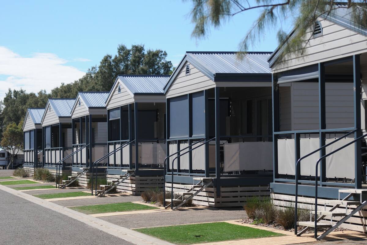 A row of modern, single-story mobile homes with external staircases, set against a backdrop of trees and a clear sky.