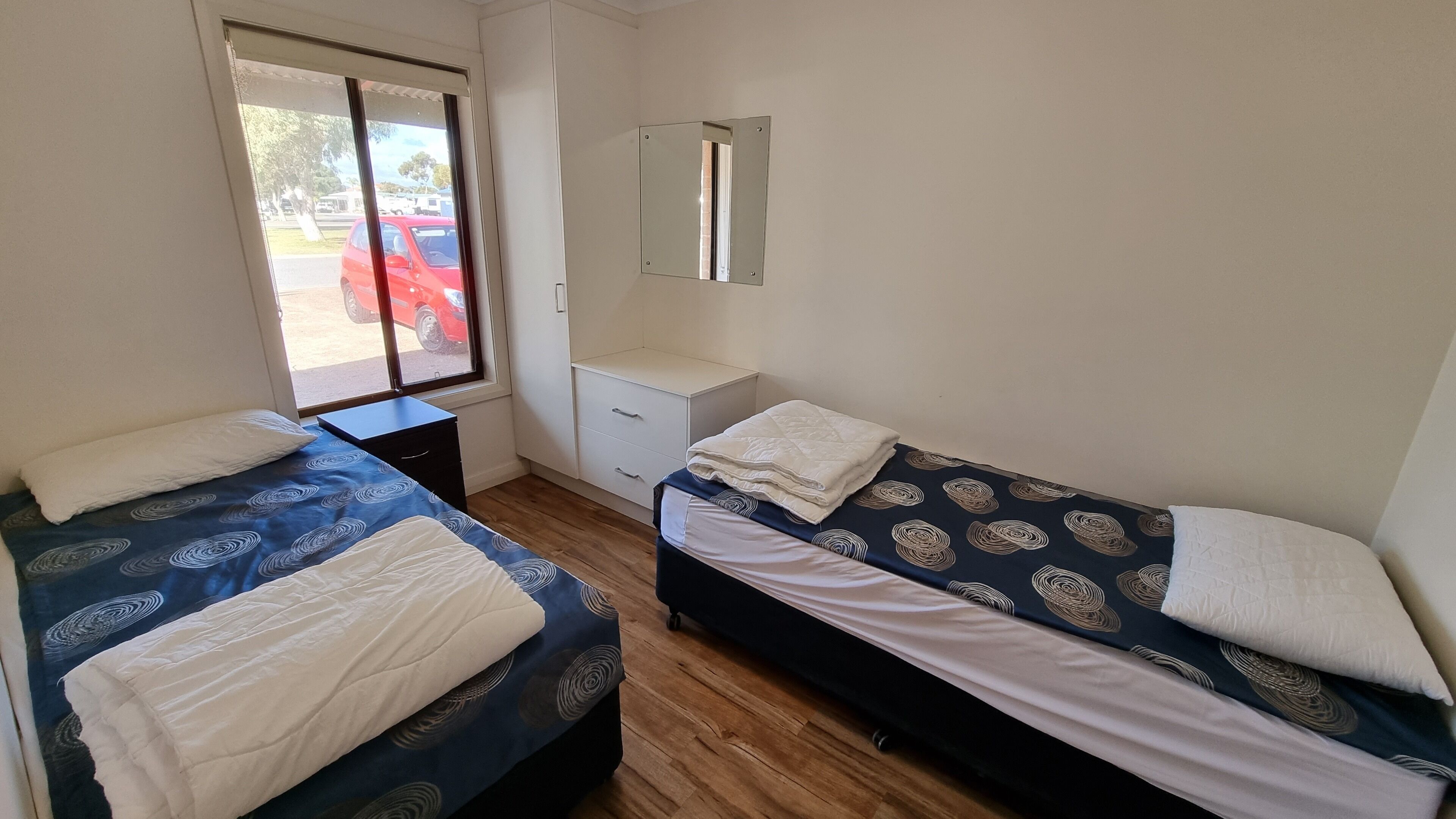 Een kamer met twee bedden, een klein bureau, een spiegel en een raam met uitzicht op een rode auto daarbuiten.