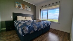 A bedroom with a large bed, a nightstand, a dresser, a window with a view of the beach, and a wall art piece. - Wallaroo Holiday Park (Wallaroo)