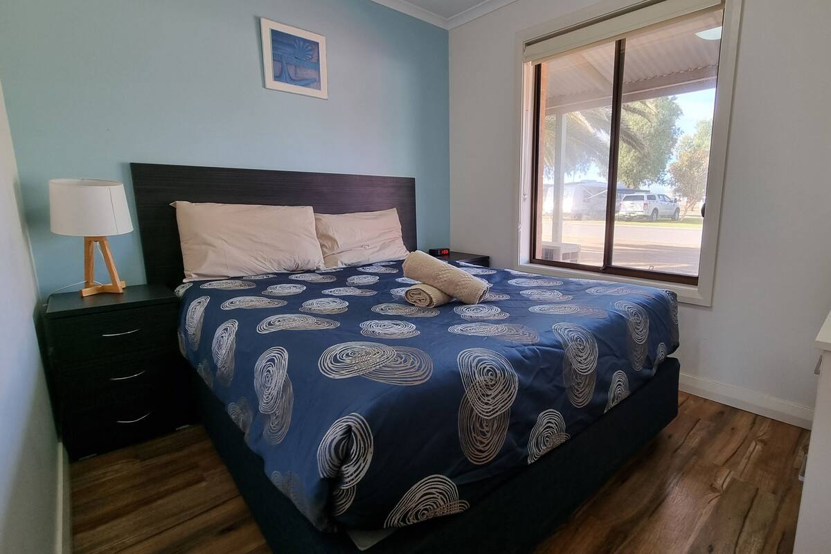 A bedroom with a bed, a nightstand, a lamp, and a window with a view of the outside.