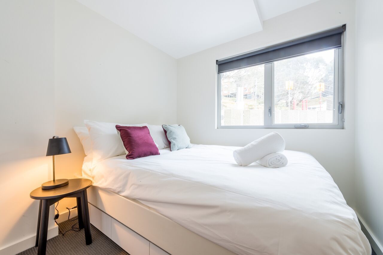 A neatly made bed with white linens and a rolled towel, a bedside lamp, and a window with a view of the outside.
