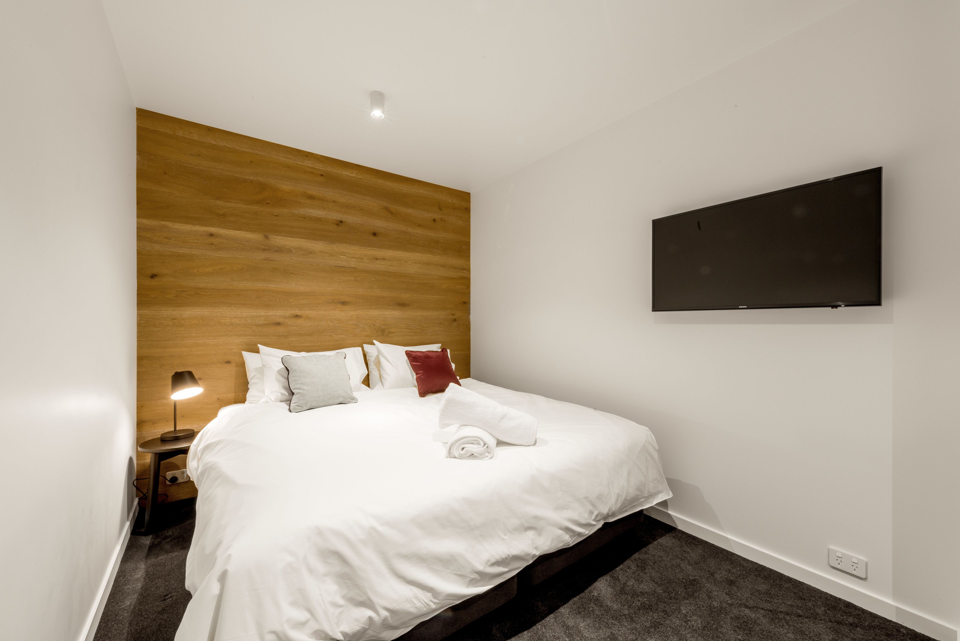 A modern hotel room with a wooden headboard, a flat-screen TV mounted on the wall, a neatly made bed with white linens, and a small bedside table with a lamp.