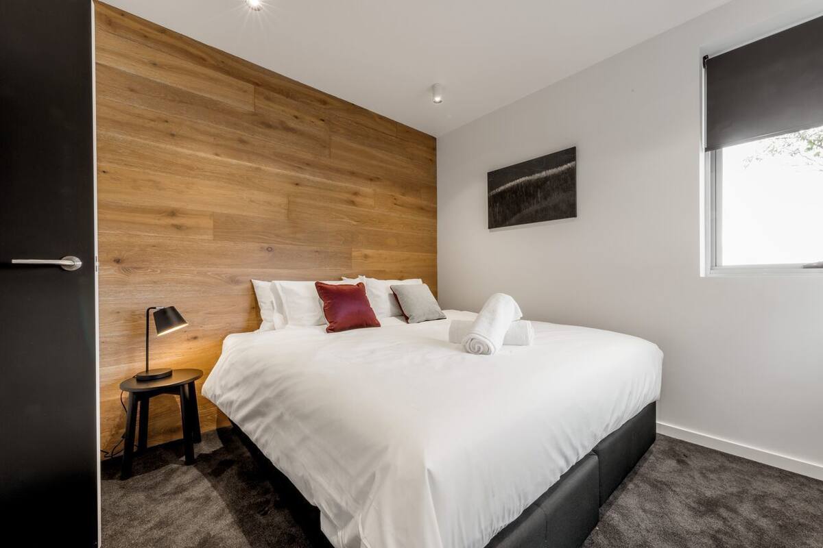 A modern bedroom with a wooden headboard, a neatly made bed with white linens, a small bedside table with a lamp, and a window with a view.
