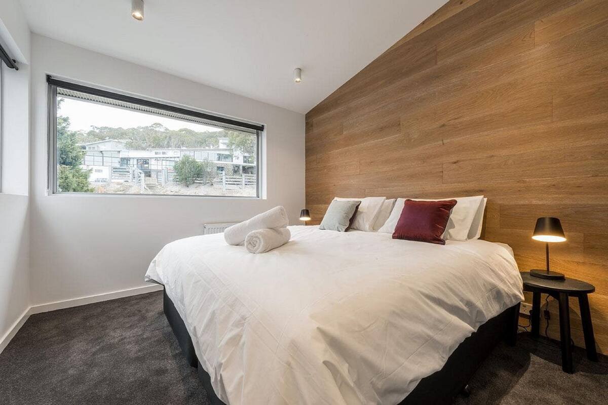 A modern bedroom with a large bed, a wooden accent wall, a window with a view, and a bedside table with a lamp.