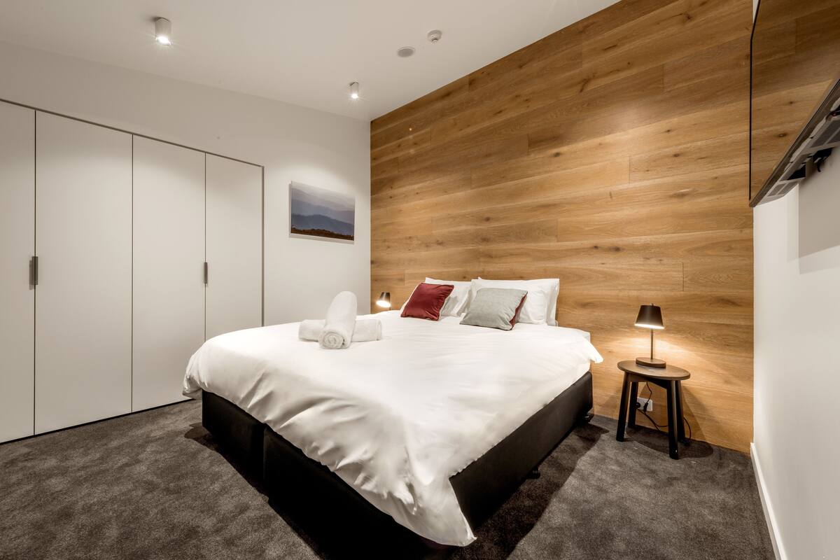 A modern bedroom with a large bed, a wooden accent wall, a small bedside table with a lamp, and a wardrobe.