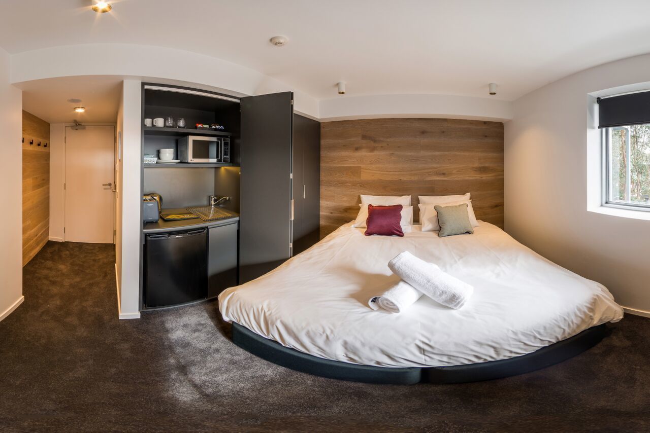 A modern bedroom with a curved bed, built-in kitchen, and a wooden headboard.