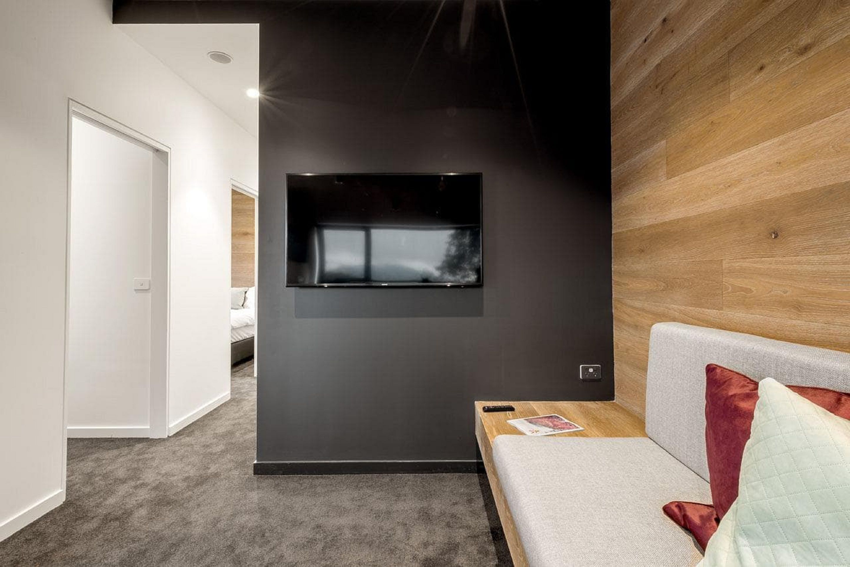 A modern bedroom with a bed, a wooden headboard, a wall-mounted TV, and a carpeted floor.