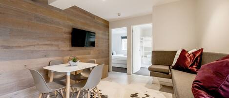 A modern living room with a wooden accent wall, a flat-screen TV, a dining area with a table and chairs, and a sofa with red cushions.