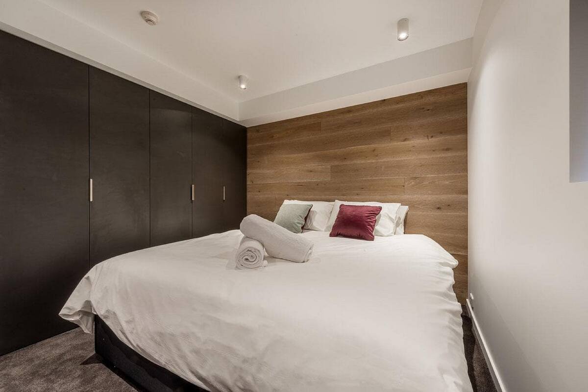 A modern bedroom with a large bed, dark wardrobe, wooden panel wall, and recessed ceiling lights.