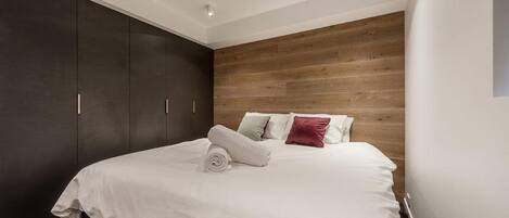 Ein modernes Schlafzimmer mit großem Bett, dunklem Wandschrank, Holzwandverkleidung und Einbauleuchten.