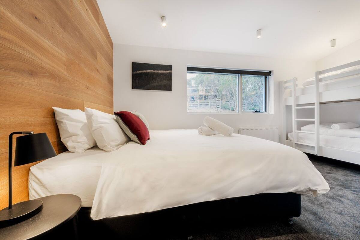 A modern hotel room with a large bed, a bedside table, a lamp, a window with a view, and built-in shelves.