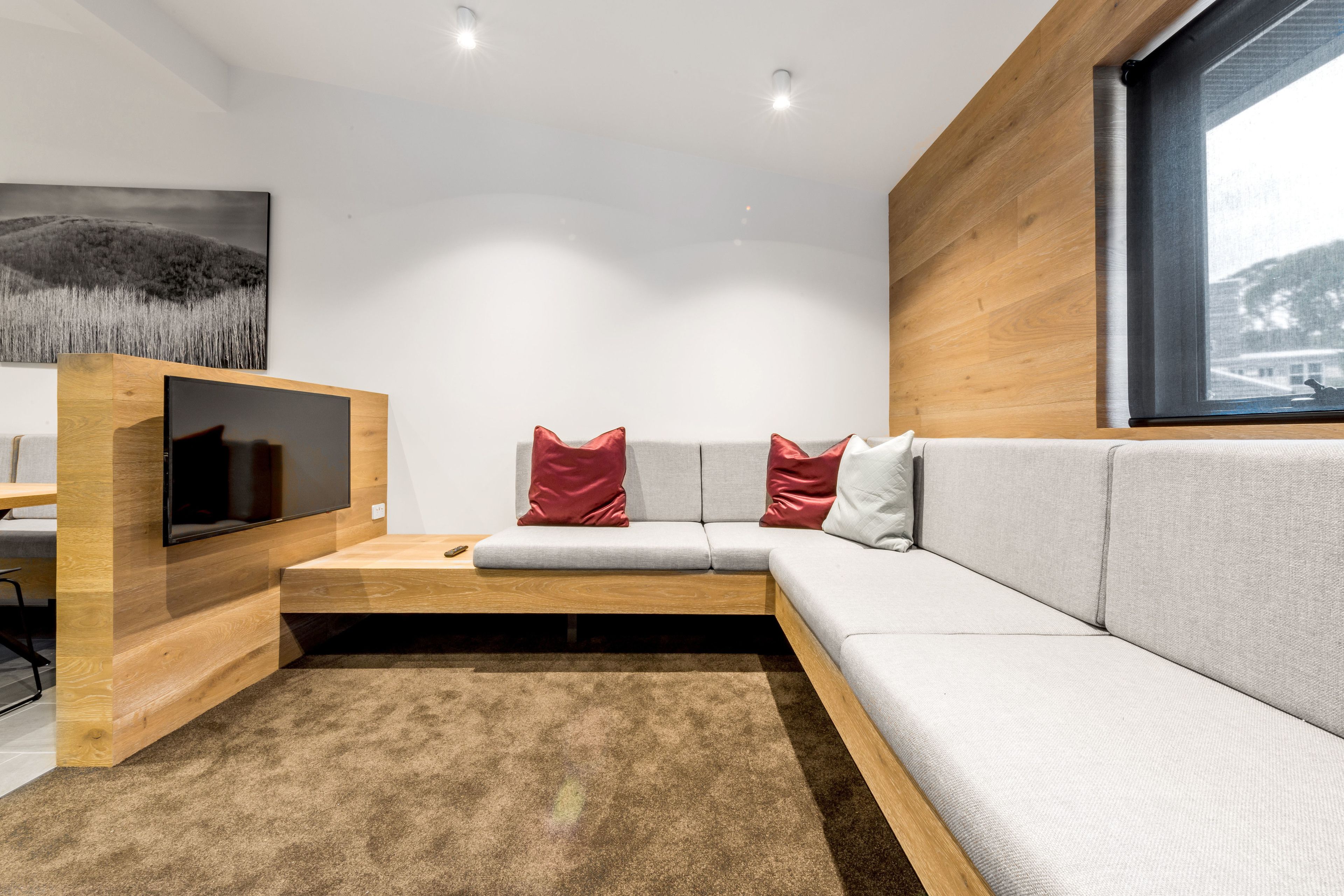 Una sala de estar moderna con un sofá gris, cojines rojos y blancos, un mueble bajo de televisión de madera y un ventanal grande.