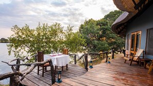 Una terraza de madera con una mesa dispuesta para comer, con vistas a un cuerpo de agua, y árboles y un edificio al fondo.