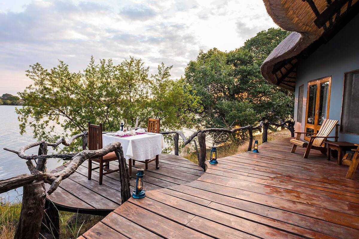 Una terraza de madera con una mesa dispuesta para comer, con vistas a un cuerpo de agua, y árboles y un edificio al fondo.