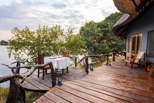 Una terraza de madera con una mesa dispuesta para comer, con vistas a un cuerpo de agua, y árboles y un edificio al fondo.