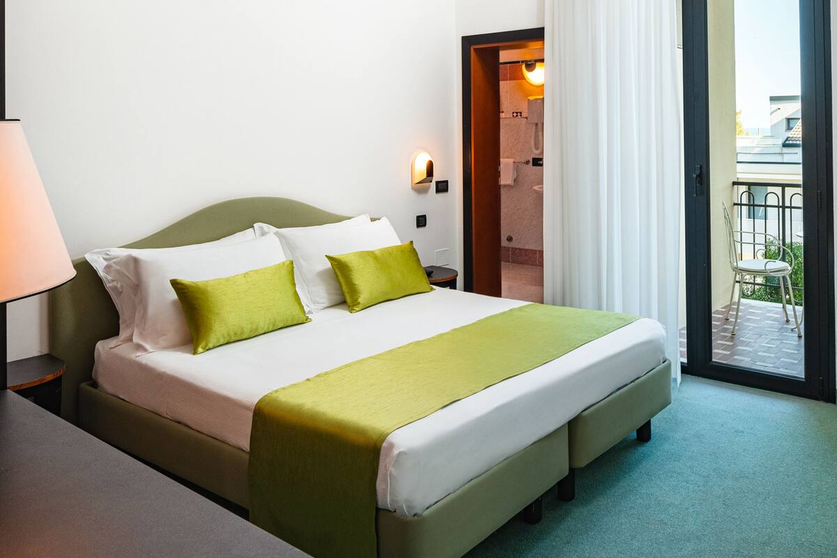 A hotel room with a bed, a nightstand, a lamp, and a view of the outside balcony.