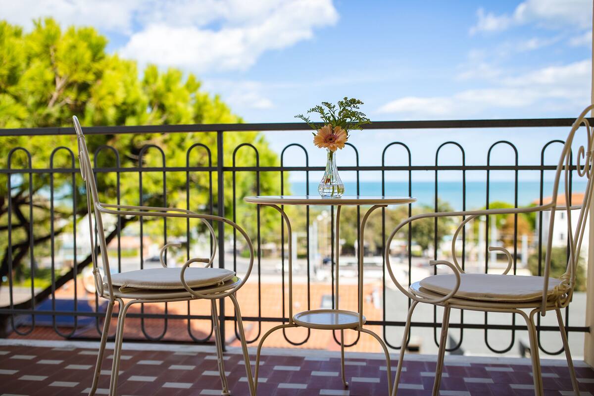 A balcony with two metal chairs and a small table, a vase with flowers, and a view of the sea.