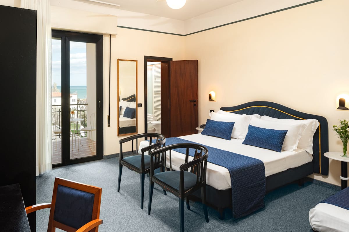 A hotel room with a bed, two chairs, a small table, a nightstand, and a view of the sea through a sliding door.