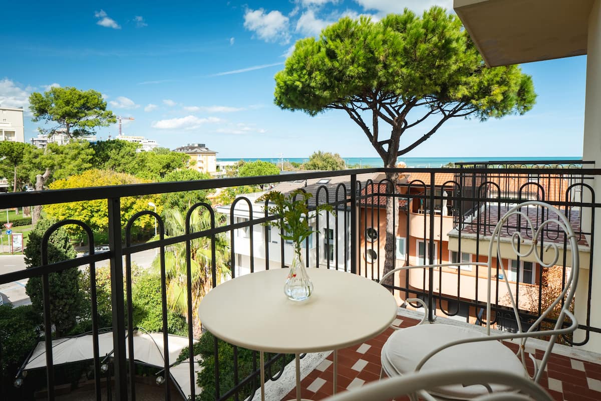 A balcony with a white table and chairs, a potted plant, and a view of the sea and buildings.