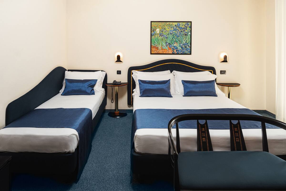 A hotel room with two beds, each with a blue headboard, white linens, and matching blue pillows. There is a small bedside table with a lamp on each side of the beds, and a painting above the headboard.