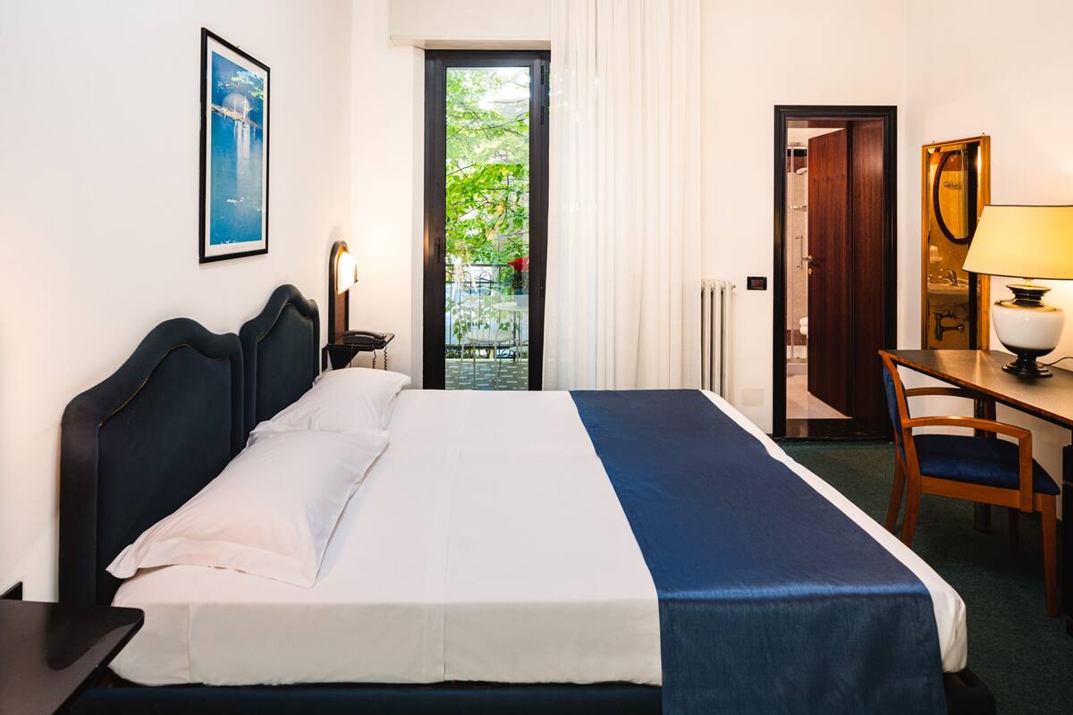 A hotel room with a bed, a desk, a chair, a lamp, and a door leading to a balcony.
