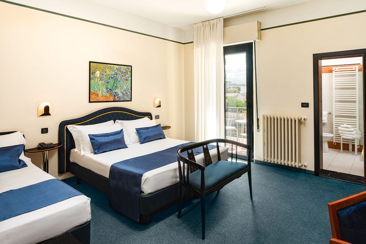 A hotel room with two beds, a chair, a painting, and a view of the outside.