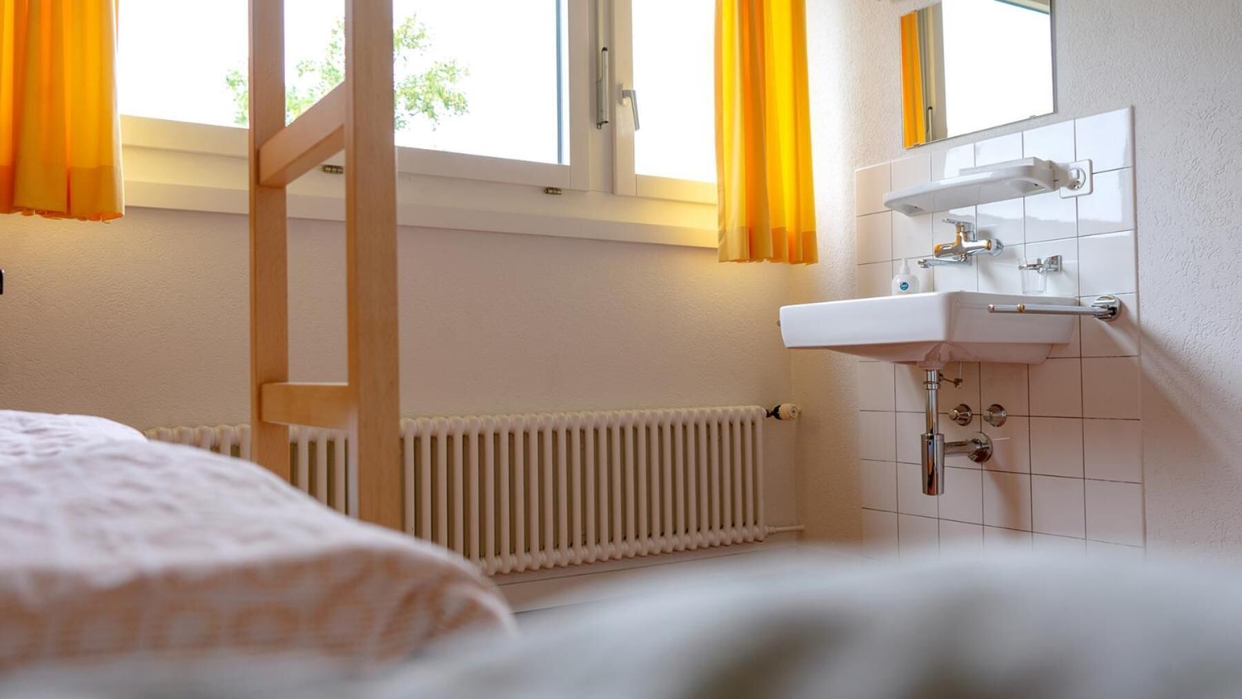 Un dormitorio con cama, ventana con cortinas, radiador y baño con lavamanos y espejo.