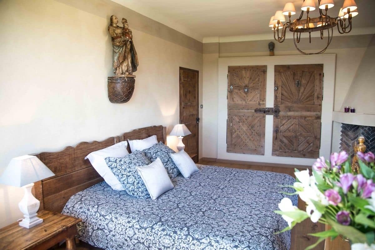 Een slaapkamer met een groot bed, een houten hoofdbord, twee nachtkastjes met lampen, een houten beeld aan de muur en een houten deur met een geometrisch patroon.