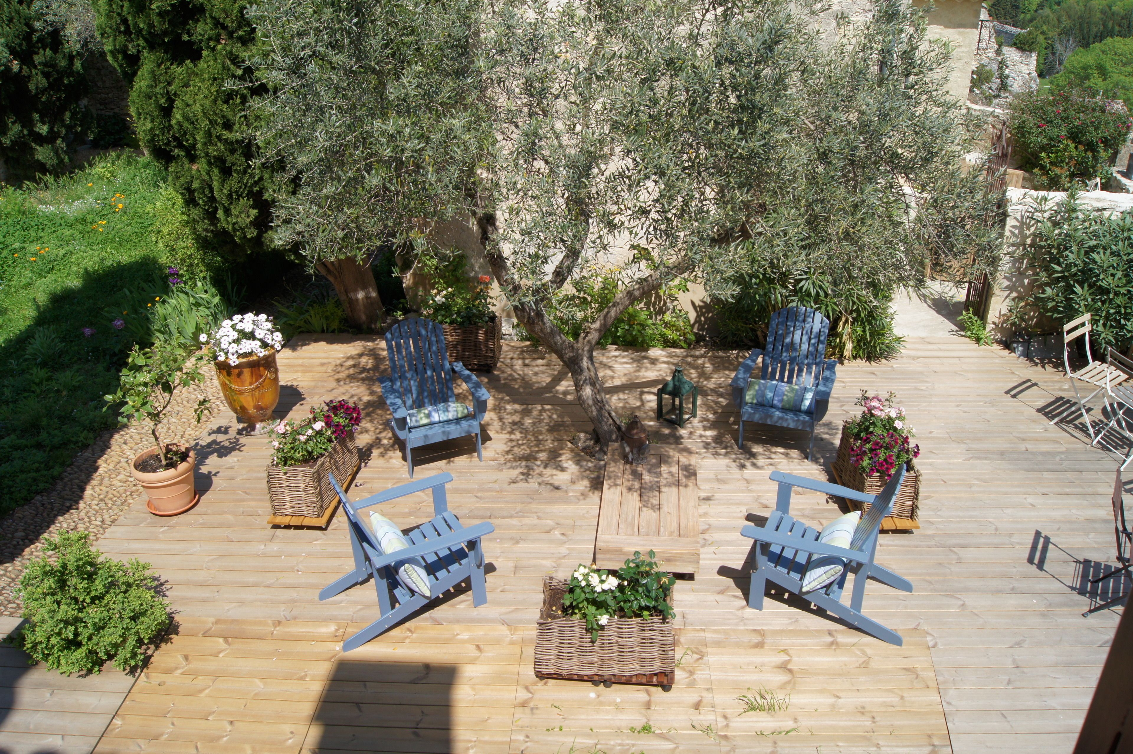 Um deck de madeira com cadeiras azuis e vasos de plantas, rodeado por árvores e vegetação.
