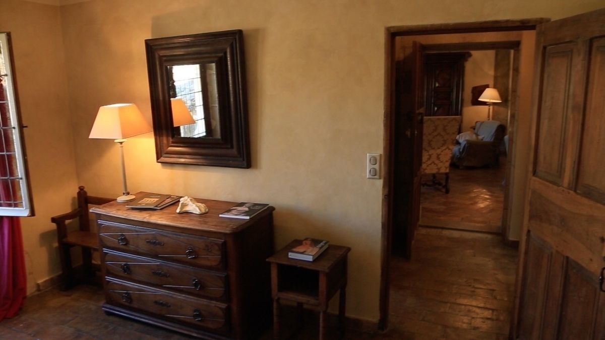 A room with a wooden dresser, a mirror, and a lamp. There is a small table with a book and a lamp. A door leads to another room with a chair and a lamp.