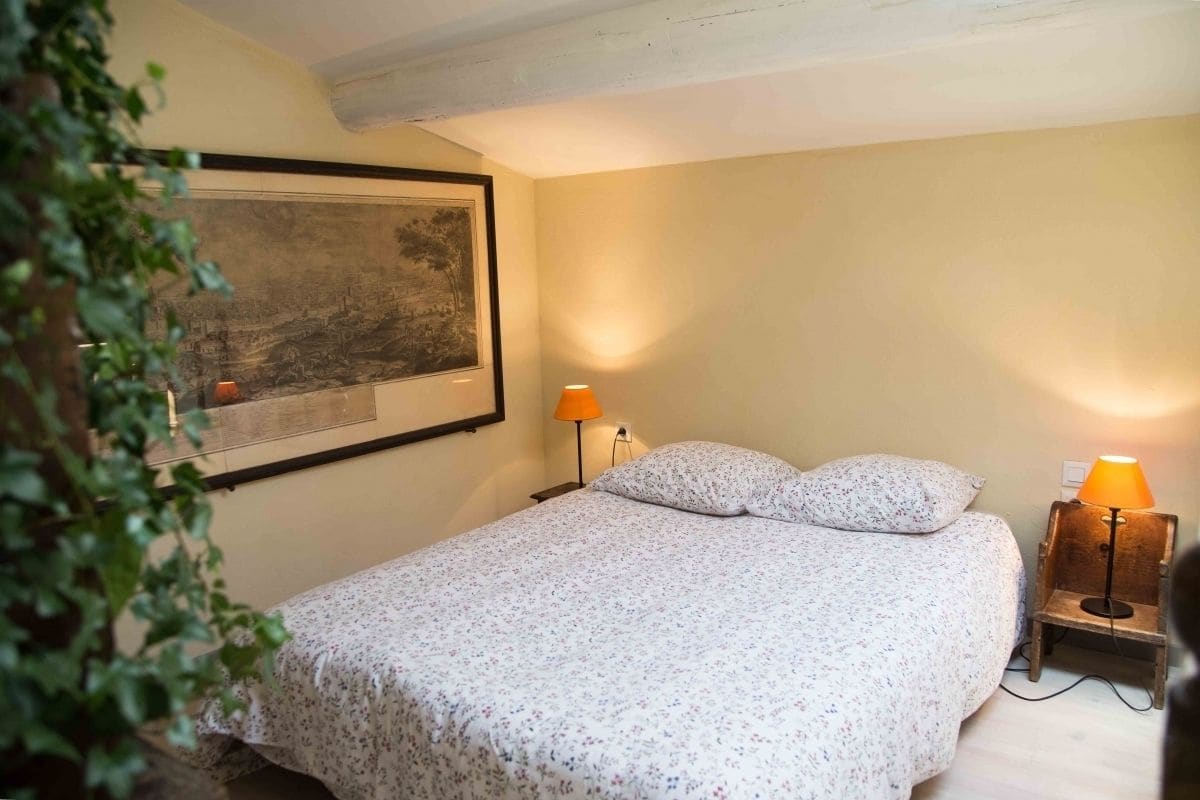 A bedroom with a bed, two bedside lamps, a framed picture on the wall, and a plant in the corner.