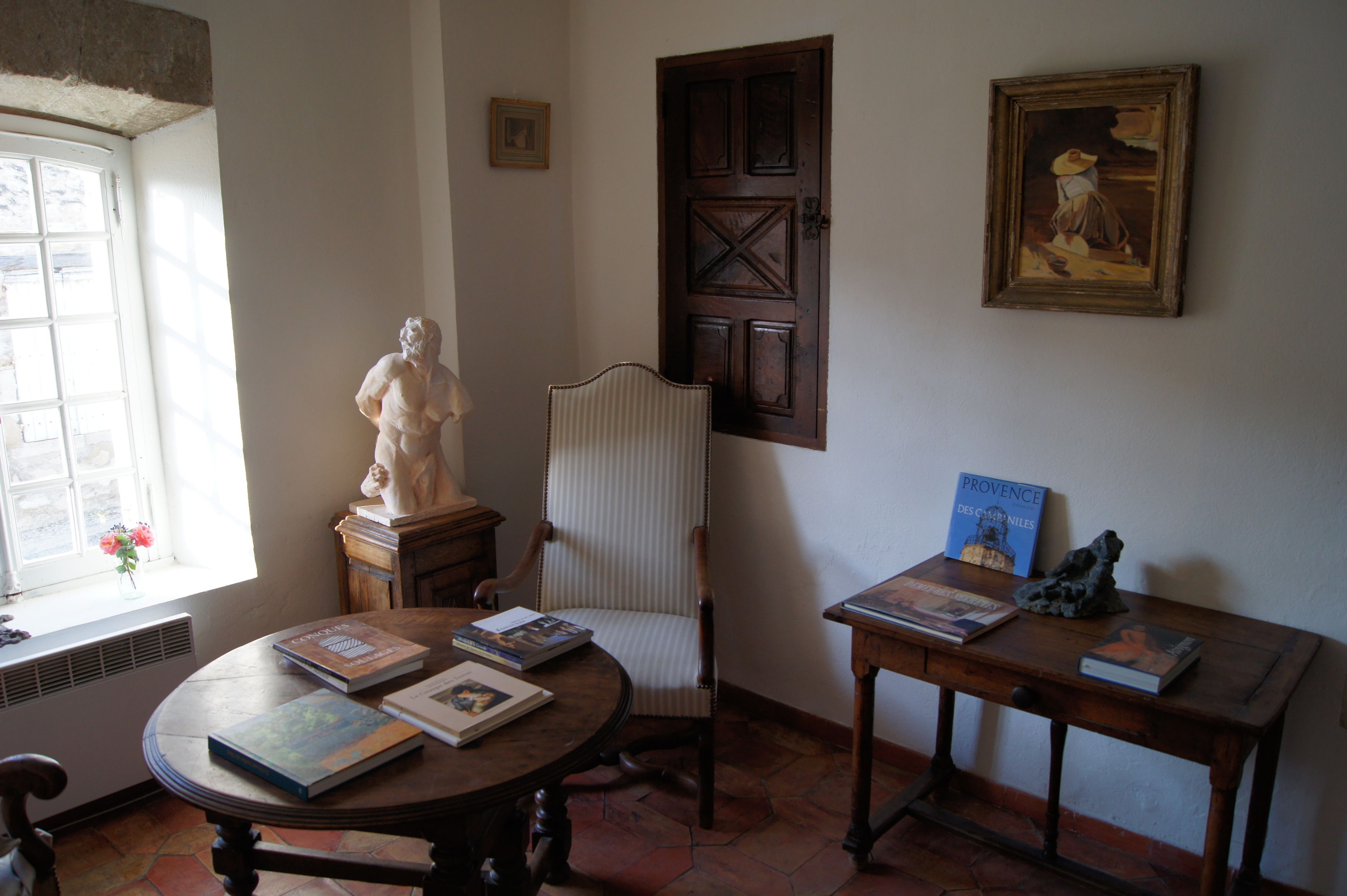 Una habitación con una mesa redonda, una silla, una estatua, un cuadro enmarcado y una ventana con flores.