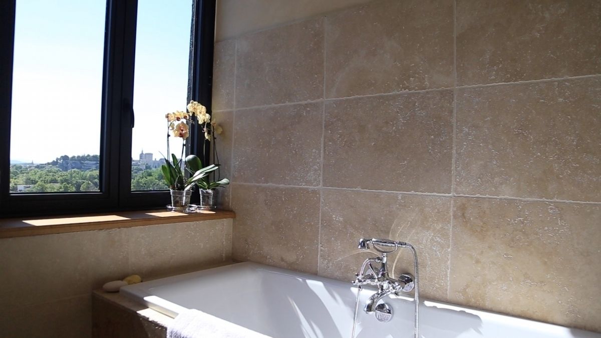 Un baño con bañera, una ventana que ofrece vistas a zonas verdes y edificios, y un florero con flores en el alféizar.