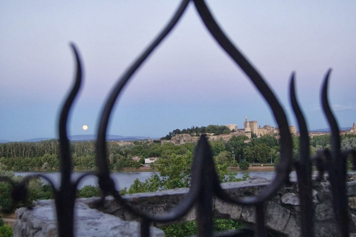 A view of a historic castle on a hill, with a river in the foreground and a fence partially obscuring the view.