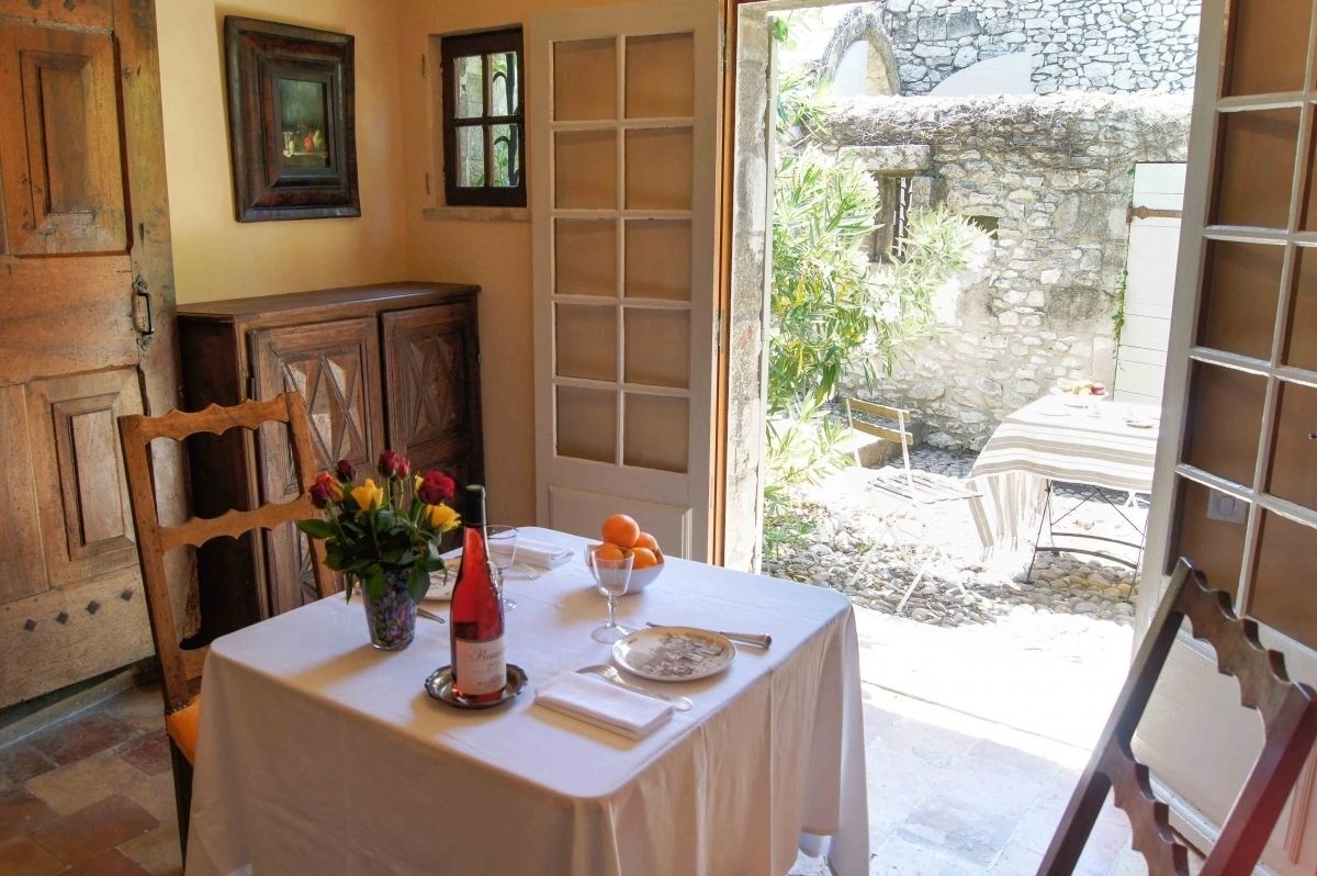 Un área de comedor con una mesa lista para dos comensales, un jarrón con flores, una botella de vino y un bol de fruta.