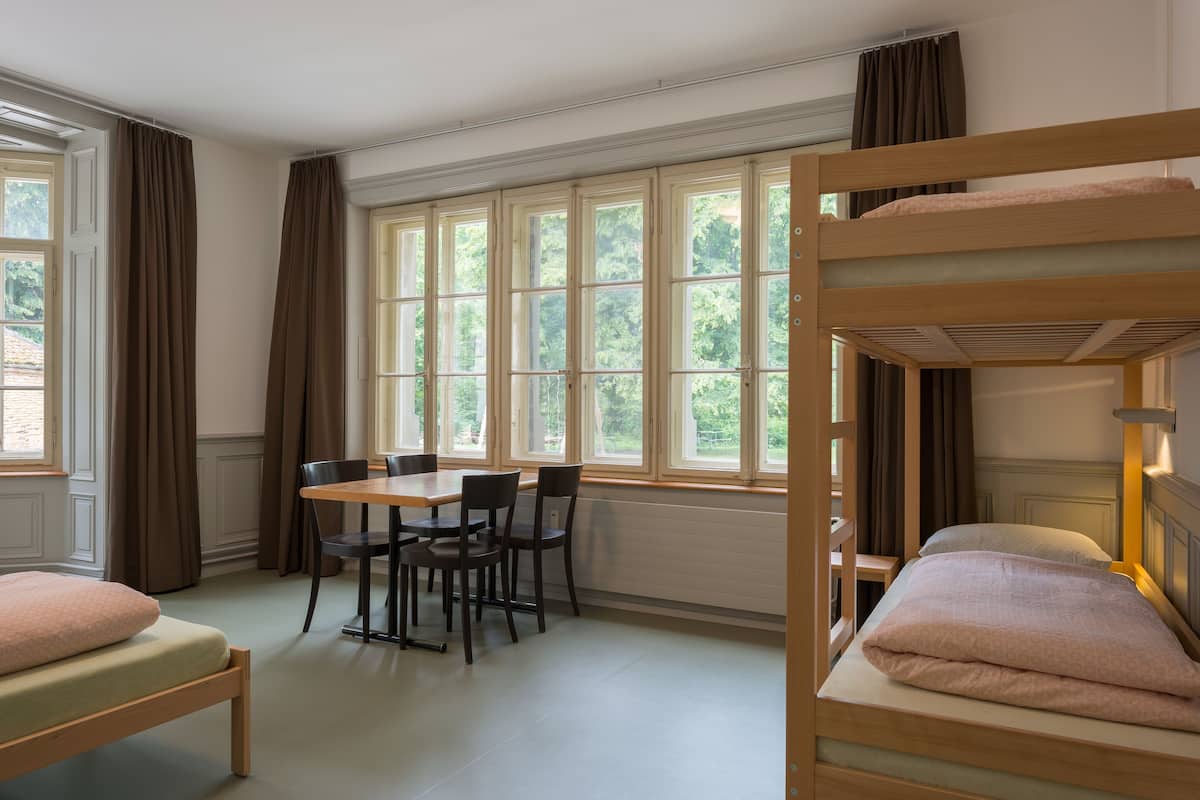 A room with bunk beds, a dining table with chairs, and large windows with curtains.