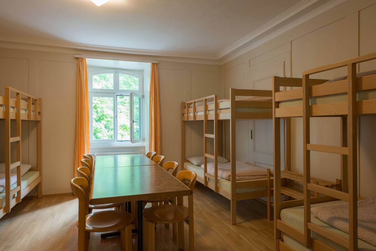 A room with bunk beds, a dining table with chairs, and a window with curtains.