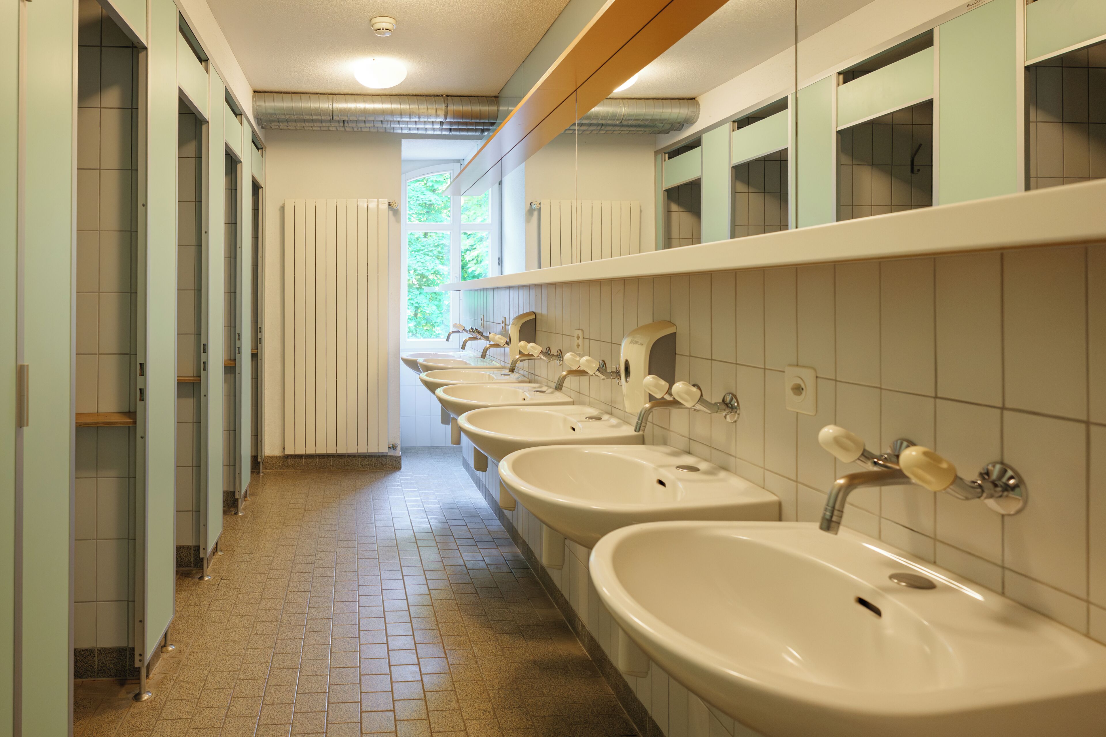 A public restroom with multiple sinks and stalls, a tiled floor, and a window providing natural light.