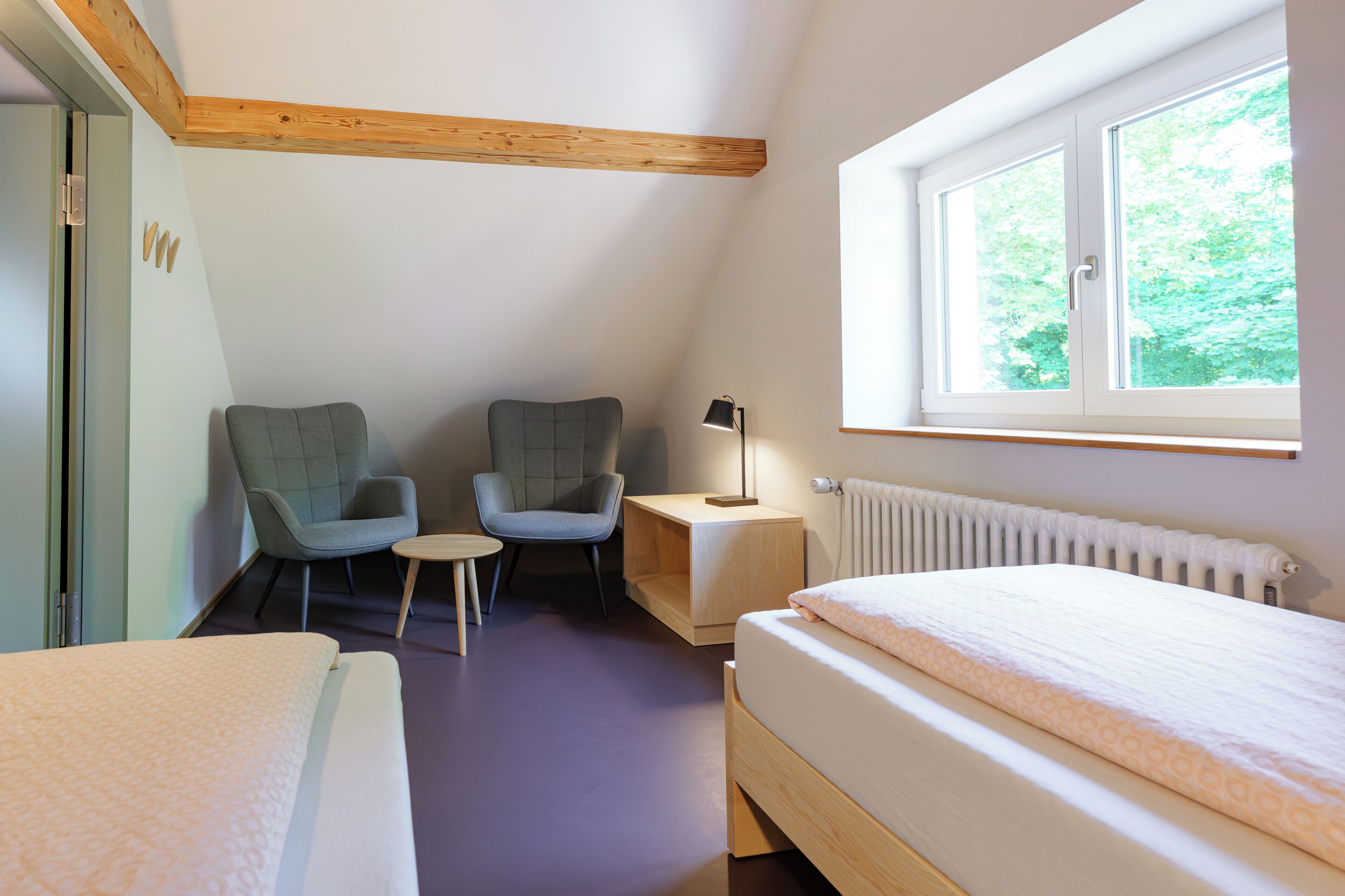 Ein Zimmer mit einem Bett, zwei Stühlen, einem kleinen Tisch, einer Lampe, einem Fenster und einer Heizung.