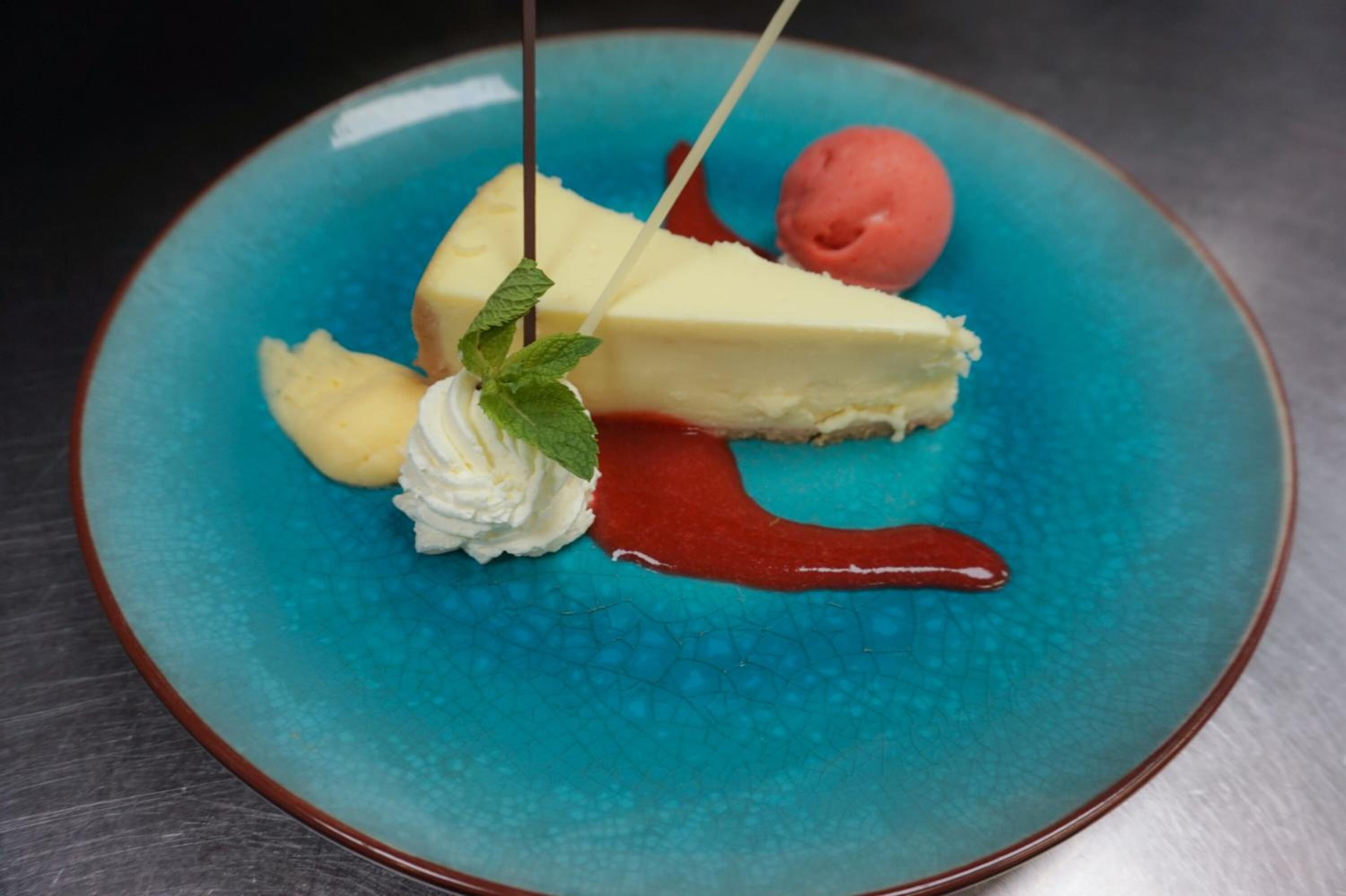A dessert plate with a slice of cheesecake, a scoop of ice cream, a strawberry, and a mint leaf on a blue textured plate.