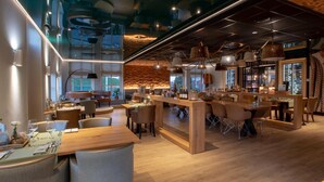 A modern restaurant with wooden tables and chairs, a bar area, and a view of the cityscape through large windows. - Hotel Termunterzijl (Termunterzijl)