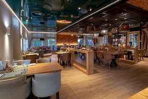 Um restaurante moderno com mesas e cadeiras de madeira, um espaço de bar e vista para a paisagem urbana através de grandes janelas.