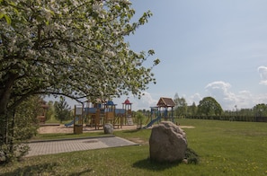 Een speelplaats met glijbanen en klimconstructies, omringd door een grasveld en bomen.