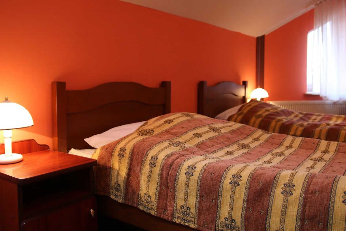 A hotel room with two beds, each with a patterned bedspread, a wooden nightstand with a lamp, and a window with curtains.
