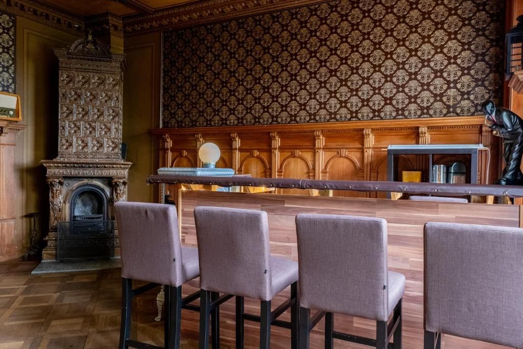 A bar area with a long wooden counter, four cushioned bar stools, a patterned wallpaper, and a statue on the right.