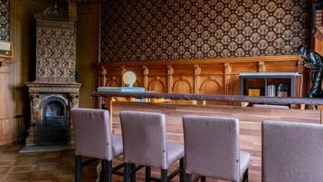 A bar area with a long wooden counter, four cushioned bar stools, a patterned wallpaper, and a statue on the right.